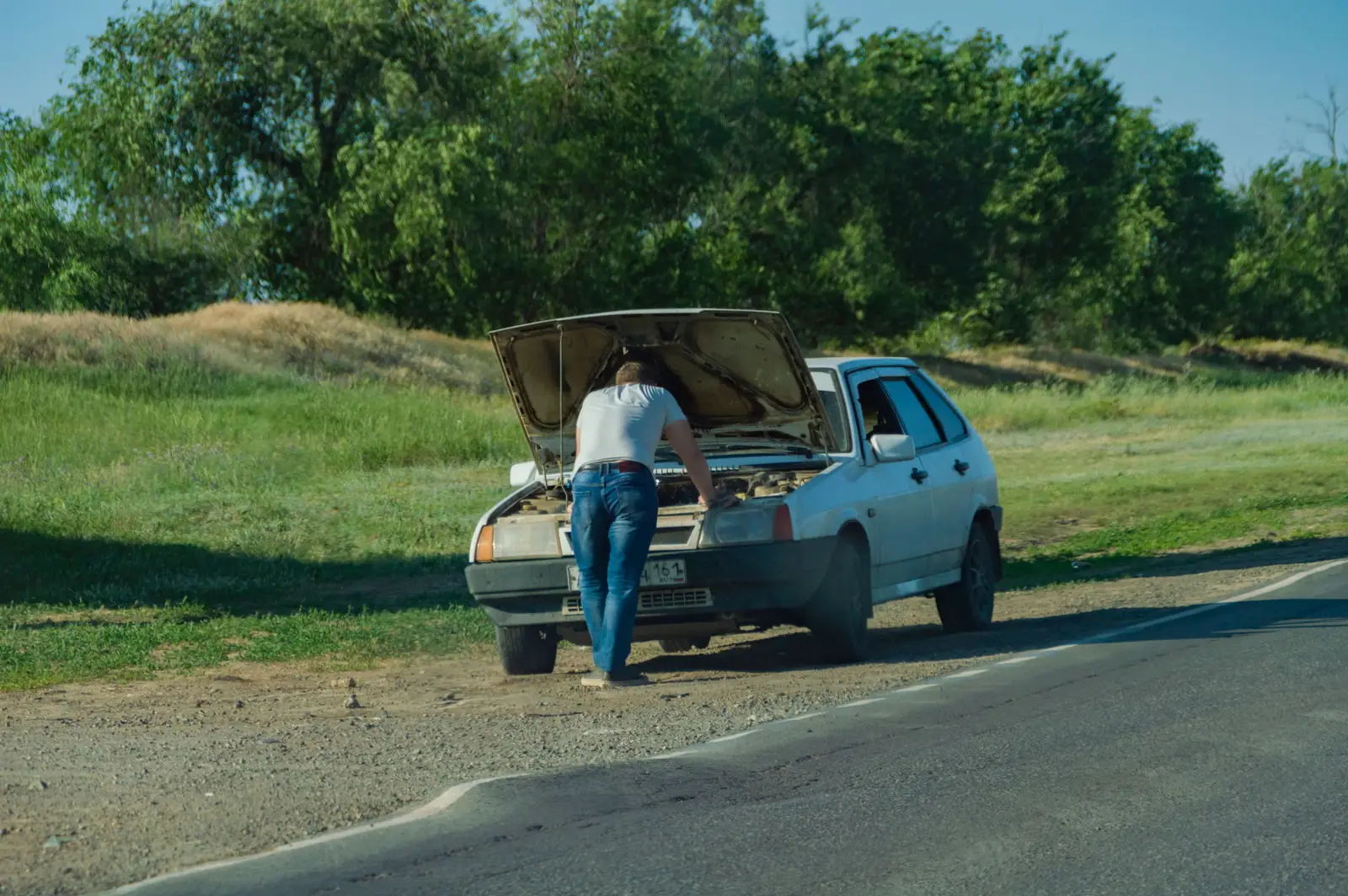 Driver looking under the raised hood of a car stopped on the shoulder of a rural road